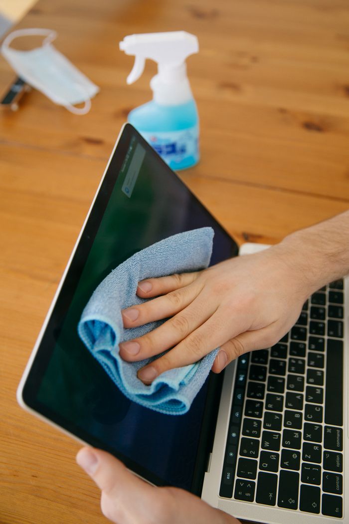 about-01 A person cleans a laptop screen with a blue cloth and spray bottle on a wooden table for hygiene purposes.